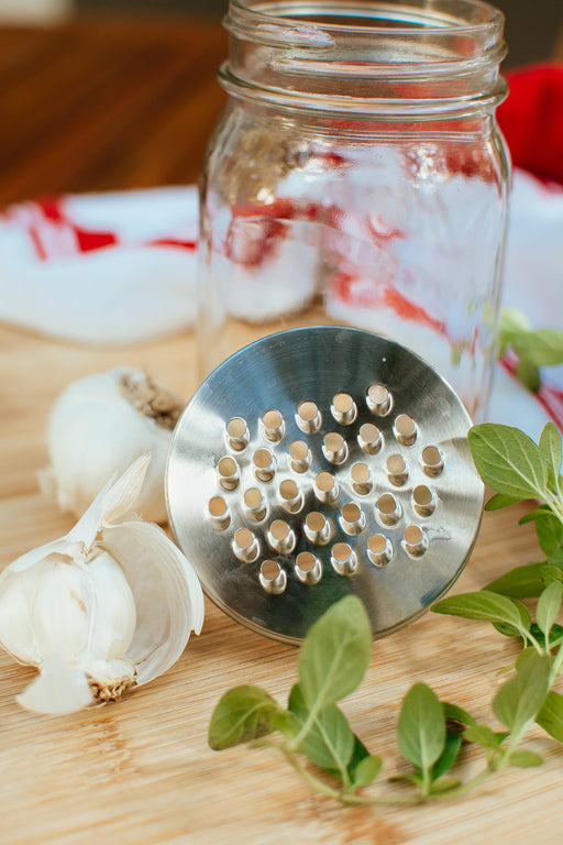 Metal garlic press on a wooden surface with garlic cloves and herbs