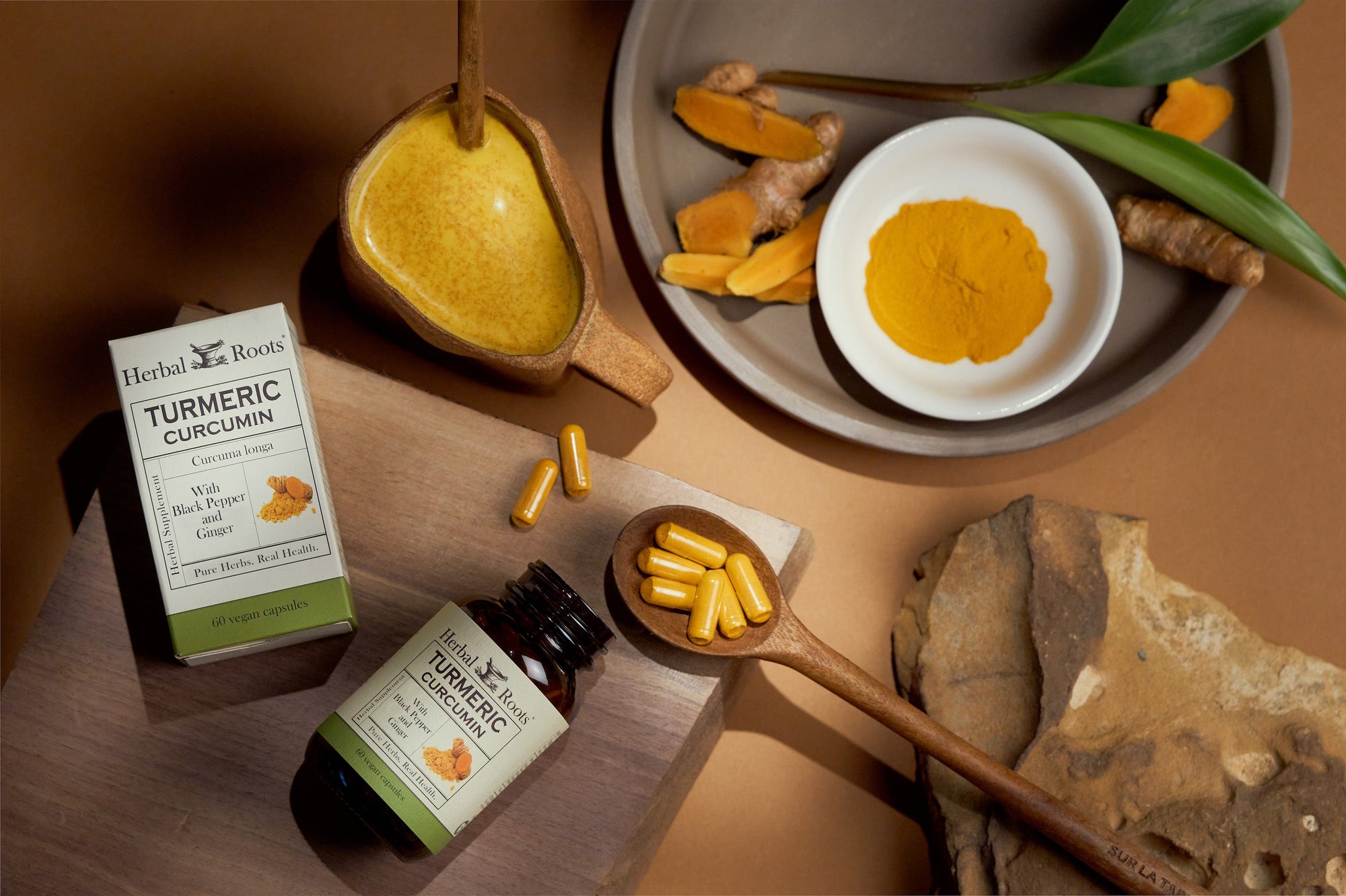 aerial view of a bottle of Herbal Roots turmeric laying on a cutting board with a wooden spoon. There are capsules in the spoon. Next to the cutting board is a mug of turmeric golden milk and a decorative plate with fresh cut turmeric root and turmeric powder.